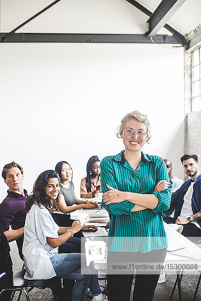 Portrait of smiling female computer hacker with arms crossed standing against team at workplace
