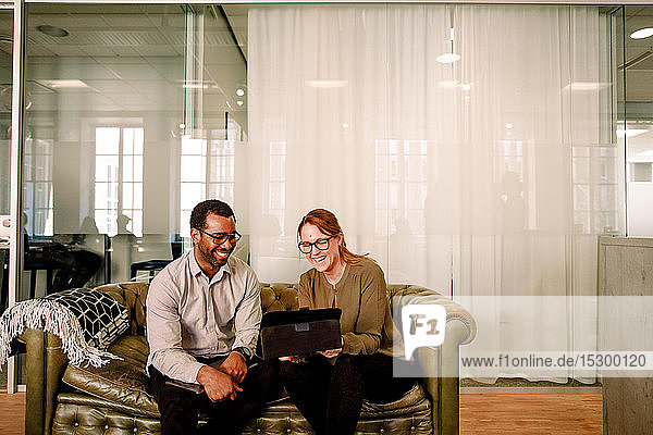Fröhliche Geschäftskollegen schauen auf das digitale Tablet  während sie im Büro auf dem Sofa sitzen