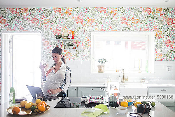 Weibliche Bloggerin benutzt Laptop  während sie ihre Tochter in der Küche zu Hause trägt
