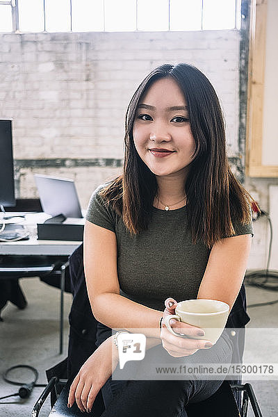 Portrait of smiling female computer programmer holding coffee cup while sitting at creative office
