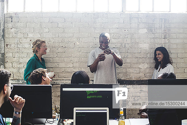 Lächelnde junge Kollegen sehen männlichen Programmierer an  der im Büro ein Smartphone an die Wand hält