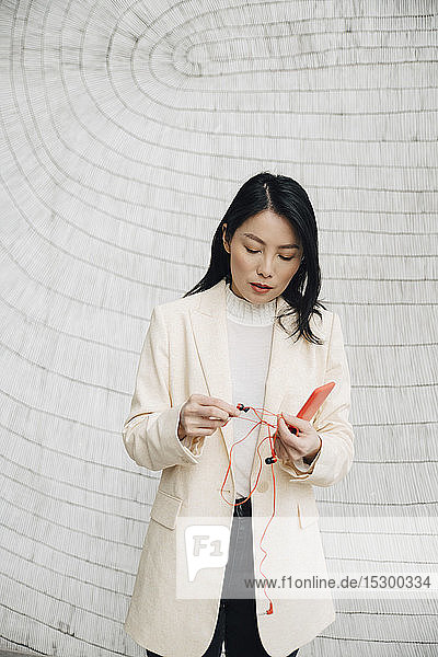 Selbstbewusste Geschäftsfrau hält Smartphone und Kopfhörer im Büro an die Wand
