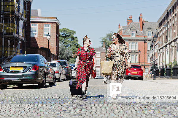Two friends walking on the street with wheeled luggage and shopping bag looking at each other Liverpool UK