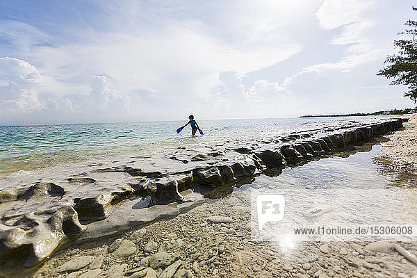5-jähriger Junge  der mit Schwimmflossen auf Felsen am Ufer läuft
