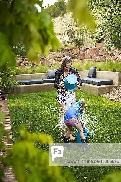 Ein junges Mädchen und ihr Bruder liefern sich im Garten eine Wasserschlacht und schütten sich gegenseitig Eimer mit Wasser aus.