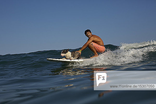 Porträt verspielter junger Mann und Hund auf dem Surfbrett auf einer Meereswelle