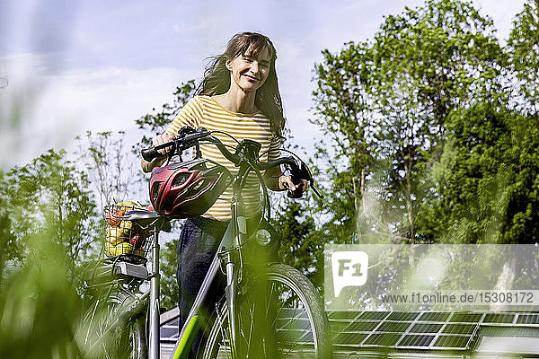 Lächelnde Frau schiebt Fahrrad mit Bio-Obst auf einer Wiese