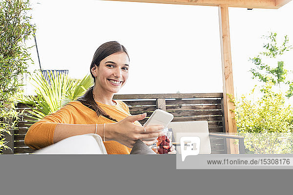 Porträt einer lächelnden jungen Frau  die mit ihrem Handy auf der Terrasse sitzt