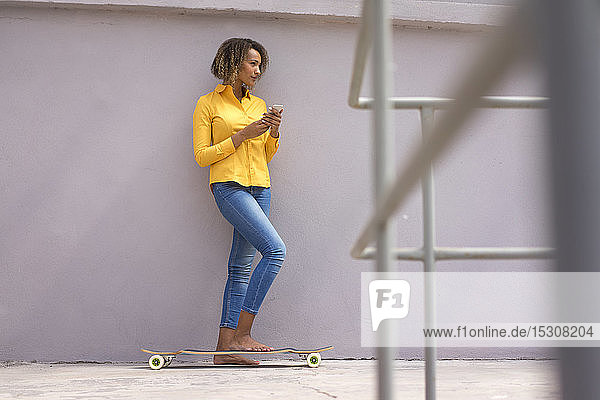 Young barefoot woman with smartphone and skateboard wearing yellow shirt and jeans