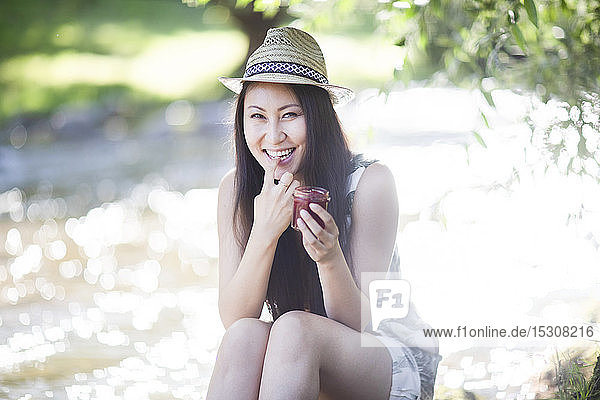 Lächelnde junge Frau mit Strohhut  die am Flussufer sitzt und knabbert