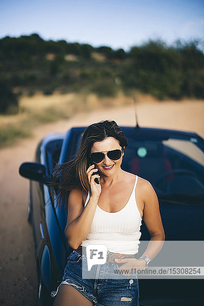 Sexy Frau  die sich an ein Cabrio lehnt und ein Smartphone benutzt