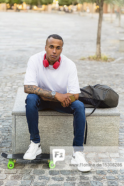 Portrait of tattooed young skateboarder sitting on bench with smartphone  headphones and backpack
