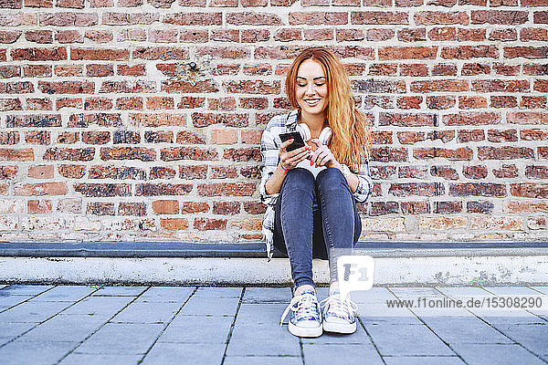 Smiling young woman sitting outdoors in the city and using smartphone