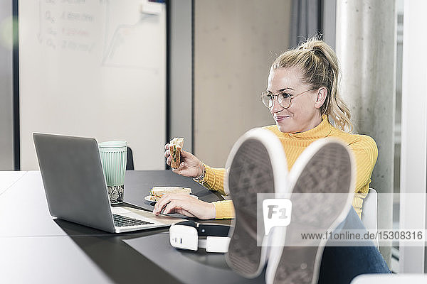 Lächelnde  ungezwungene Geschäftsfrau  die im Büro am Tisch sitzt  ihren Laptop benutzt und ein Sandwich isst