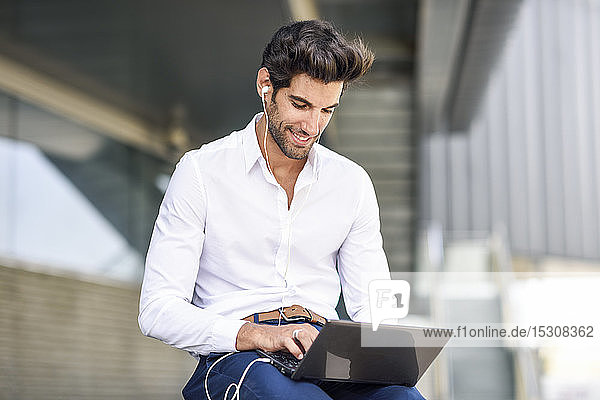 Smiling businessman wearing earphones using laptop outdoors in the city