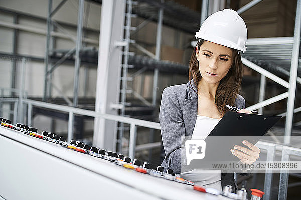 Woman wearing hard hat at control panel in a factory taking notes