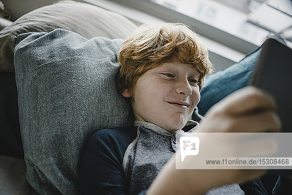 Porträt eines lächelnden rothaarigen Jungen  der mit Hilfe eines digitalen Tablets auf der Couch liegt