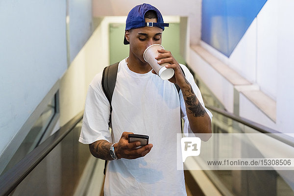 Tätowierter junger Mann mit Rucksack steht auf Rolltreppe und schaut auf Handy  während er Kaffee zum Mitnehmen trinkt