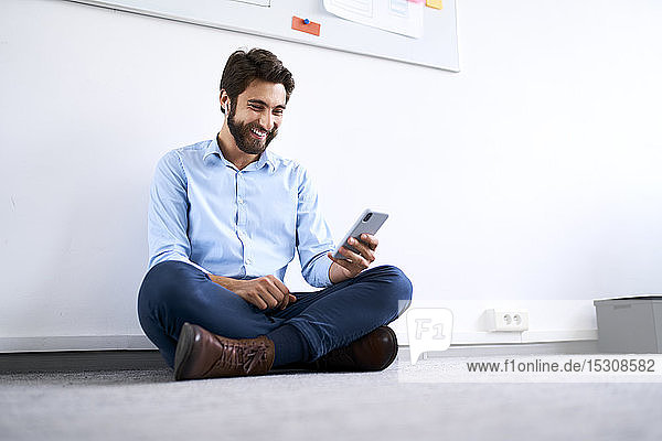 Lächelnder Geschäftsmann  der auf dem Boden sitzt und sein Smartphone benutzt