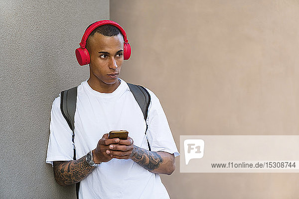 Porträt eines tätowierten jungen Mannes  der mit Smartphone und roten Kopfhörern Musik hört