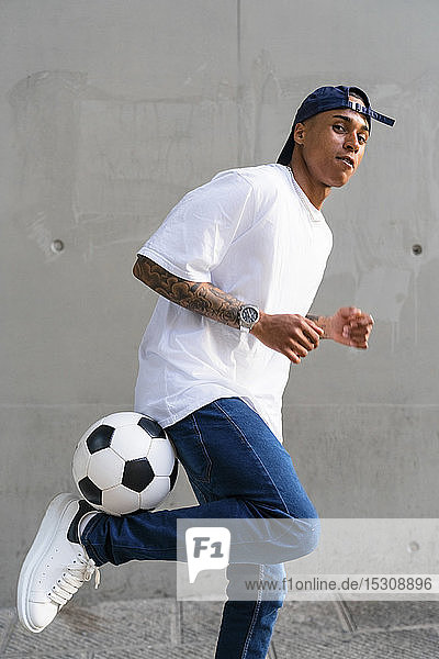 Porträt eines tätowierten jungen Mannes  der vor einer Betonwand Fußball balanciert