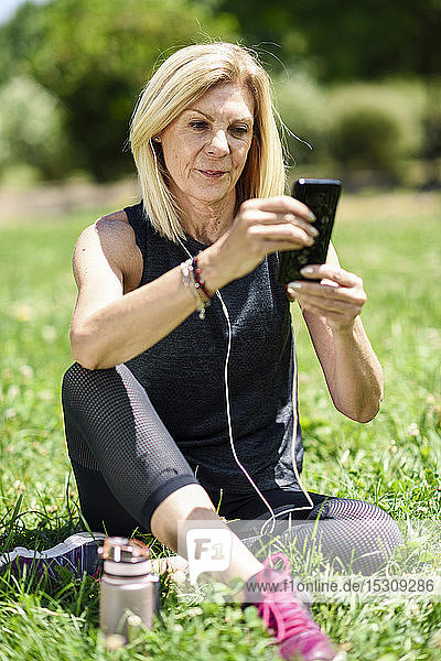 Reife Frau  die ihr Smartphone benutzt und sich nach dem Sport im Gras eines Parks ausruht