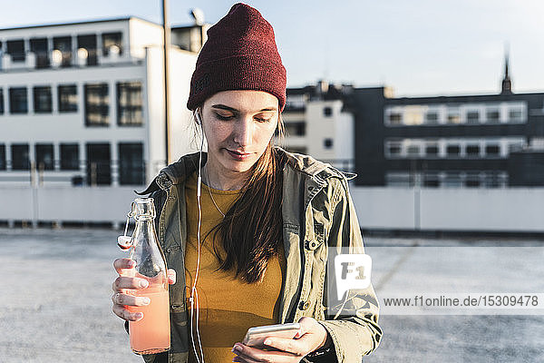 Stilvolle junge Frau mit Getränk  Kopfhörern und Mobiltelefon auf dem Parkdeck