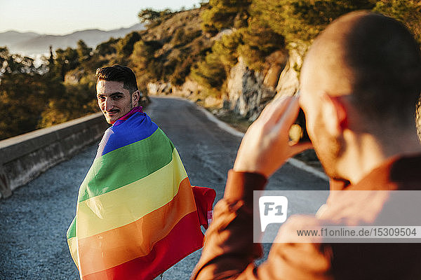 Fotograf beim Fotografieren eines Mannes  der auf einer Straße in den Bergen in eine Fahne aus schwulem Stolz gehüllt ist