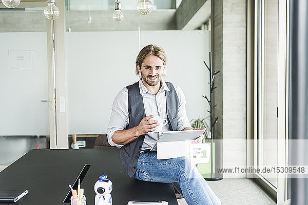 Portrait of smiling young businessman sitting on table in office with tablet and cup of coffee