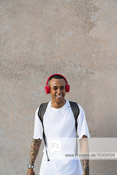 Porträt eines lachenden jungen Mannes mit Rucksack  der mit roten Kopfhörern Musik hört