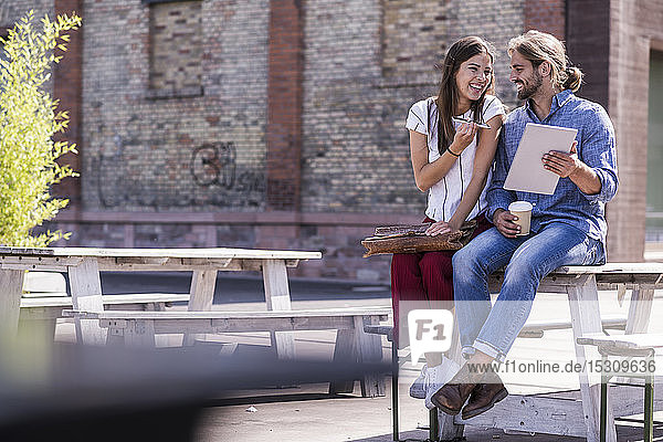 Glückliches junges Paar sitzt mit Handy und Tablet am Tisch in einem Biergarten