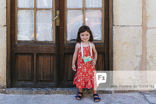 Porträt eines lächelnden kleinen Mädchens mit Holzspielzeugkamera in rotem Kleid mit Blumenmuster