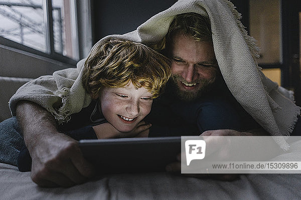 Vater und Sohn liegen zusammen unter einer Decke und schauen auf ein digitales Tablett