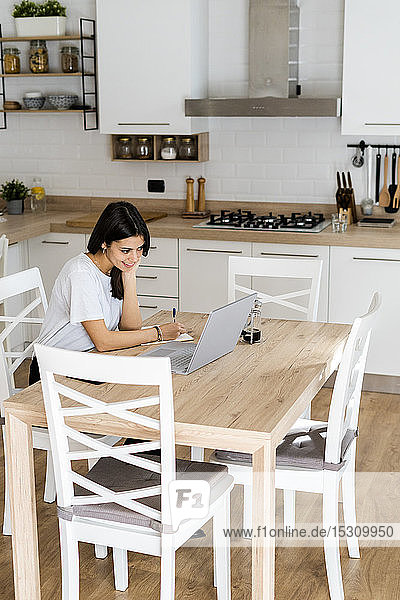Young woman with laptop taking notes at home