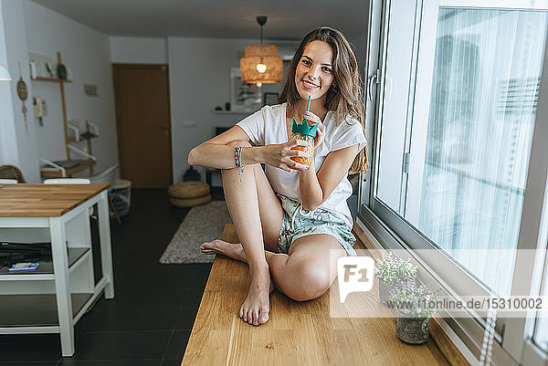 Porträt einer lächelnden jungen Frau  die auf der Küchentheke sitzt und etwas trinkt