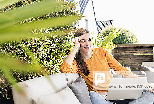 Porträt einer lächelnden jungen Frau  die mit Laptop auf der Terrasse sitzt