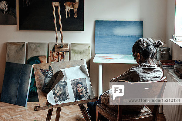 Rückansicht einer Künstlerin  die in ihrem Atelier vor einem Gemälde sitzt