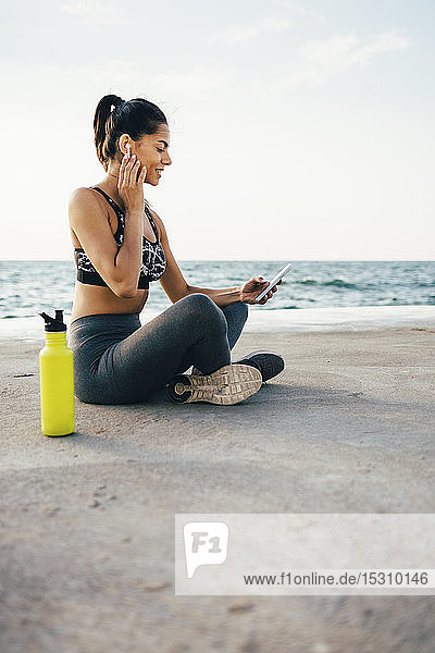 Frau benutzt Smartphone und In-Ohr-Gerät während des Trainings und sitzt auf einem Pier