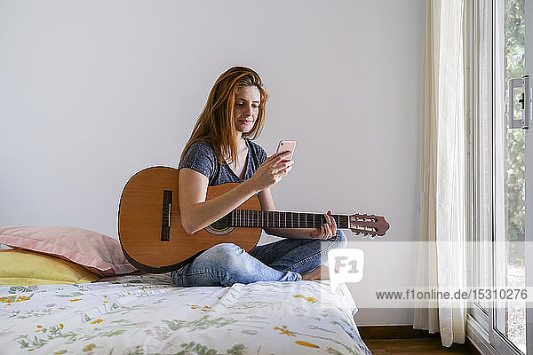 Junge Frau zu Hause chillt mit Gitarre im Schlafzimmer und benutzt ihr Smartphone