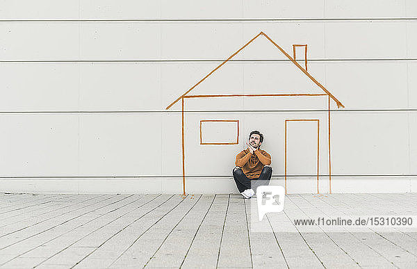 Digitale Komposition eines jungen Mannes  der mit einem Haus an einer Wand sitzt