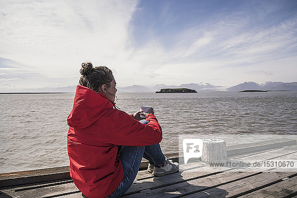 Junge Frau sitzt auf einem Steg und trinkt Kaffee  Südost-Island