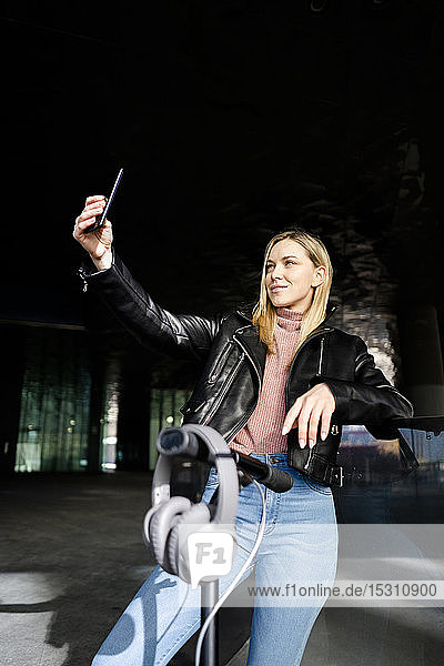 Junge Frau mit Kickroller und Kopfhörern nimmt Selfie mit Smartphone
