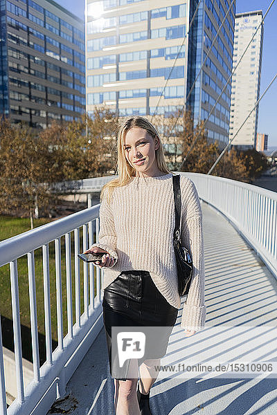 Porträt einer blonden jungen Frau mit Smartphone  die auf einer Fußgängerbrücke geht