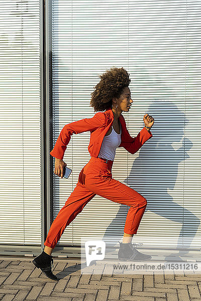 Laufende junge Frau mit Smartphone in modischem roten Hosenanzug
