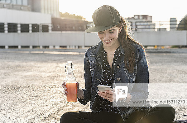 Lächelnde junge Frau mit Getränk und Mobiltelefon  die bei Sonnenuntergang auf dem Parkdeck sitzt