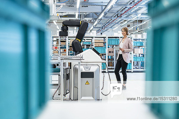 Geschäftsfrau mit Tablette in einer modernen Fabrikhalle mit Blick auf Roboter