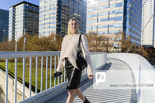 Porträt einer blonden jungen Frau mit Smartphone  die auf einer Fußgängerbrücke geht