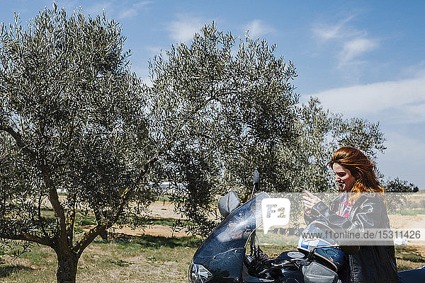 Lächelnde rothaarige Frau auf Motorrad mit Blick aufs Handy  Andalusien  Spanien