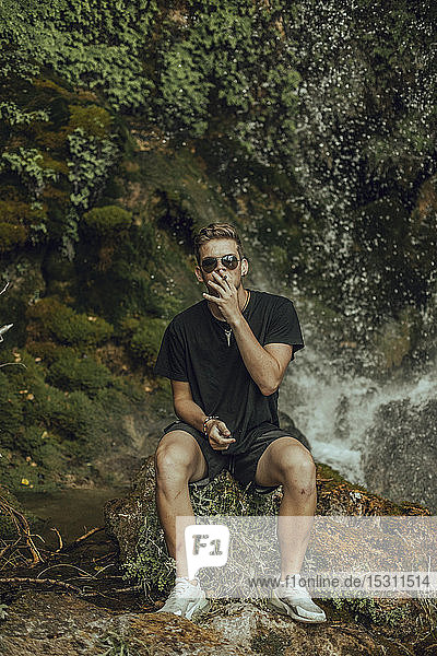 Porträt eines coolen jungen Mannes an einem Wasserfall  der einen Joint Marihuana raucht