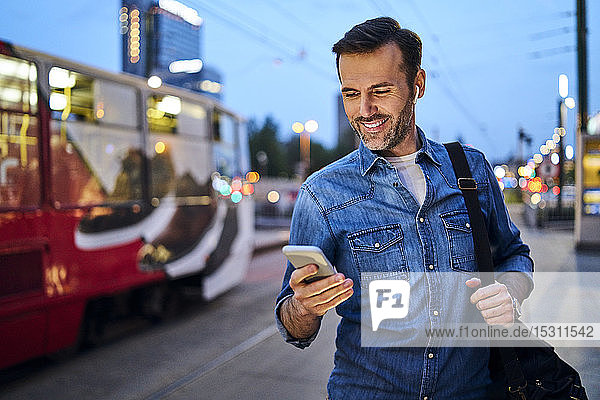 Mann benutzt Smartphone und hört Musik  während er abends an der Straßenbahnhaltestelle steht
